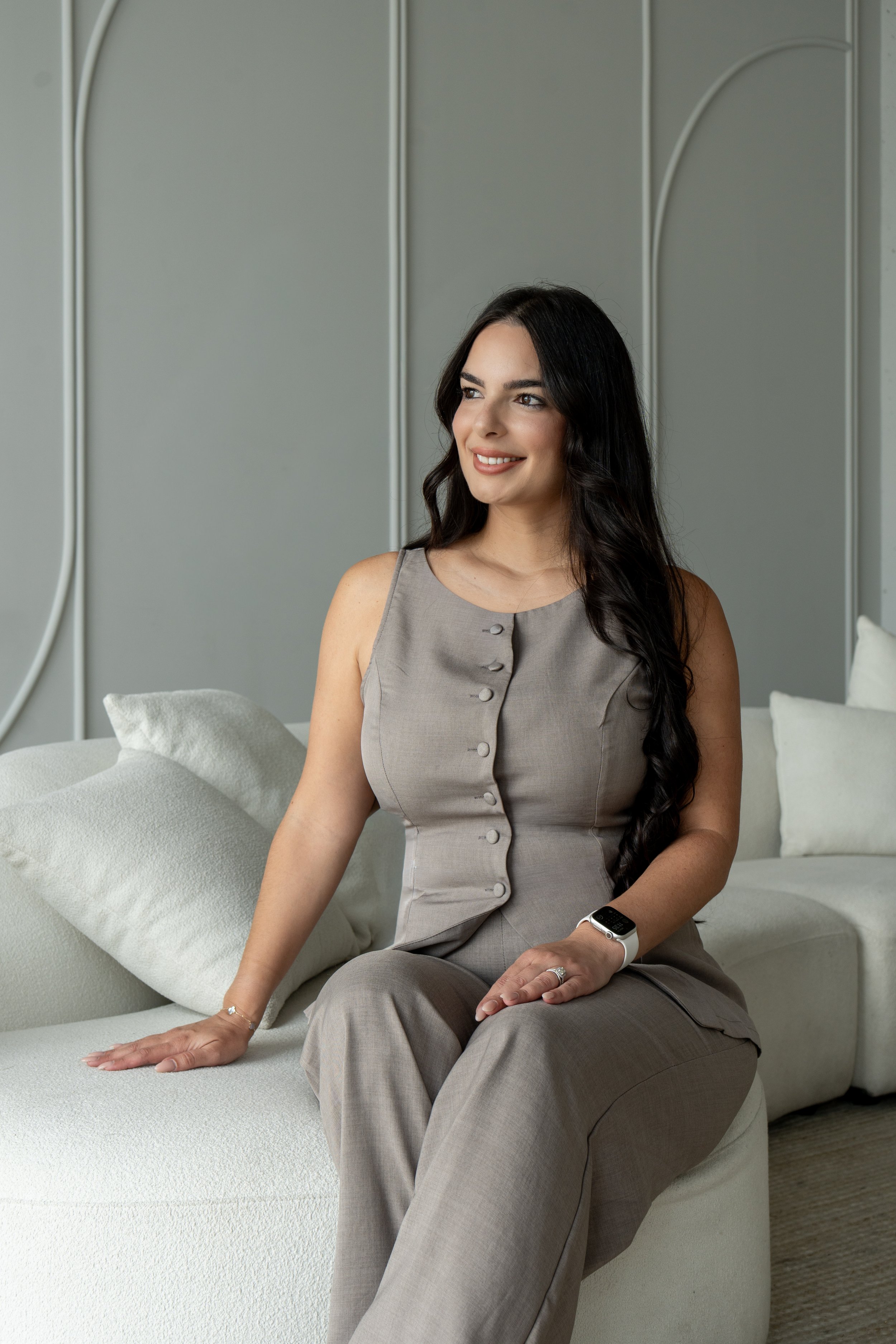 A smiling woman with long dark hair sitting on a white sofa in a modern living room, wearing a sleeveless beige top and matching pants, with a smartwatch on her left wrist.