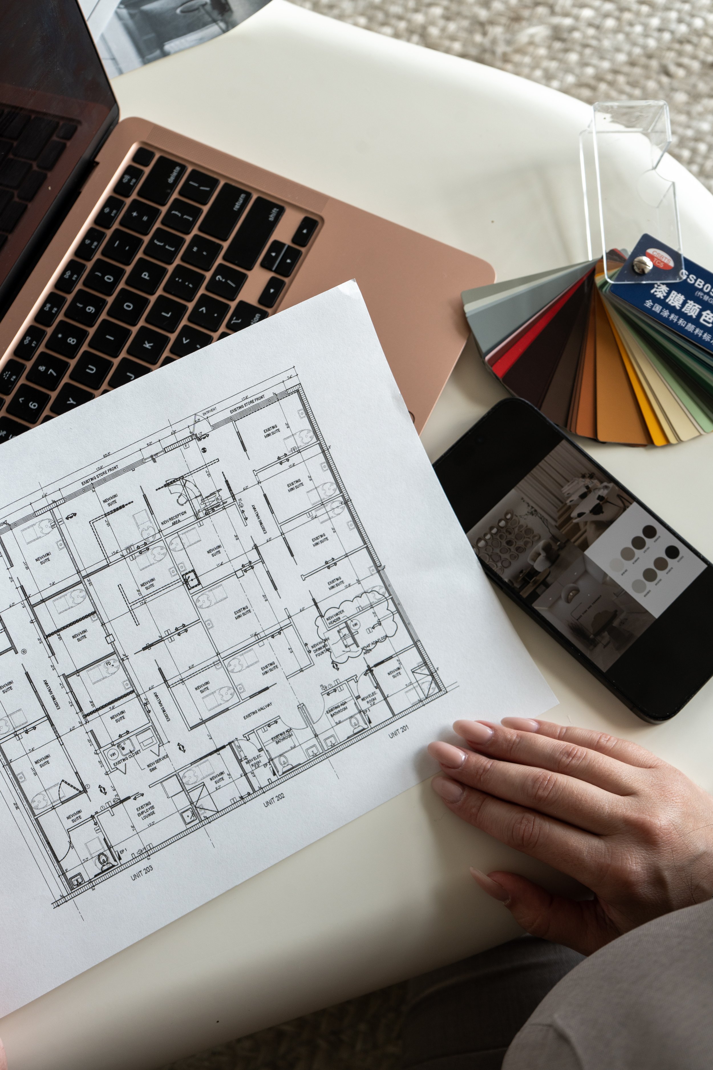 A person's hand resting on an architectural blueprint of a building layout, with a laptop, color swatches, a smartphone, and a book nearby on a white table.