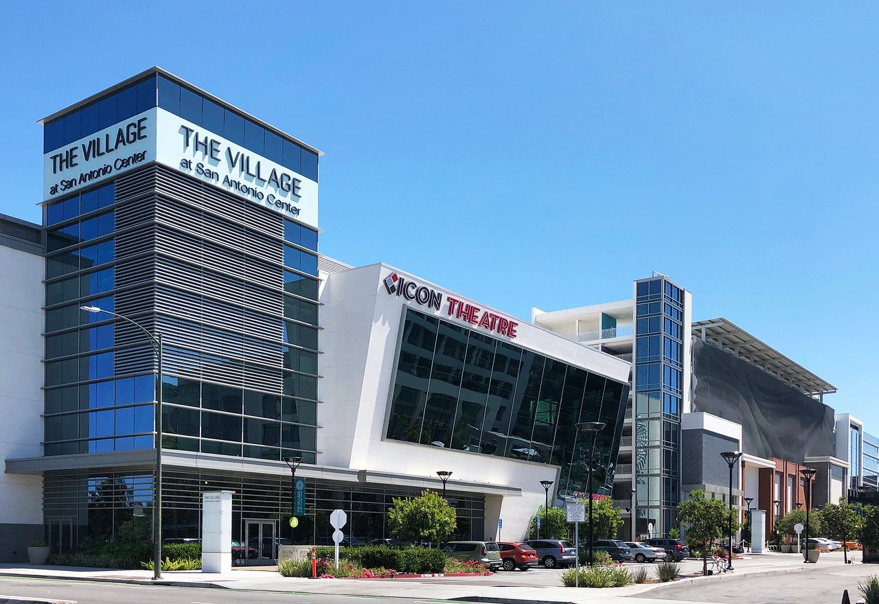Modern multi-story building with signage for The Village at San Antonio Center and ICON Theatres, with a parking lot and street view in bright daylight.