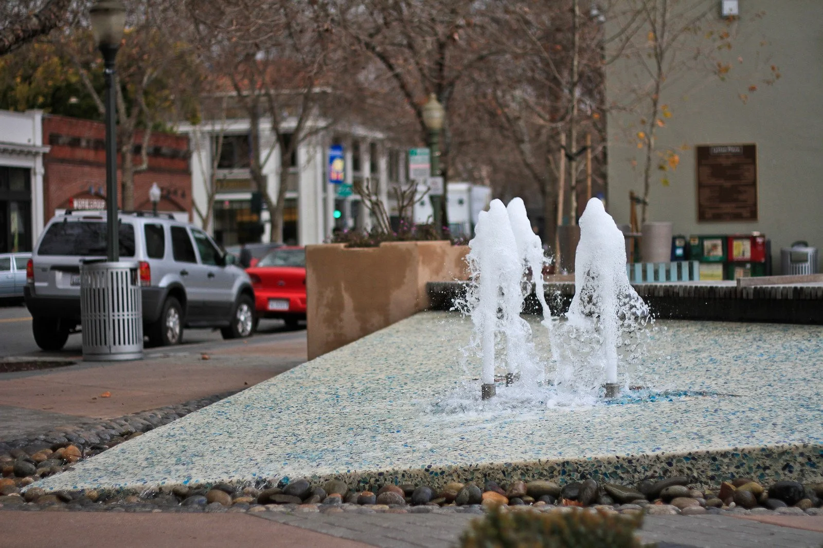 A small urban water fountain with multiple jets of water spray in a city sidewalk area, surrounded by pebbles and a concrete surface, with cars and buildings in the background.