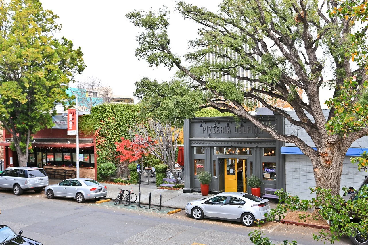 Street scene featuring a restaurant called Pizzeria Delfina with a yellow door, trees, parked cars, and bicycles, in an urban setting.