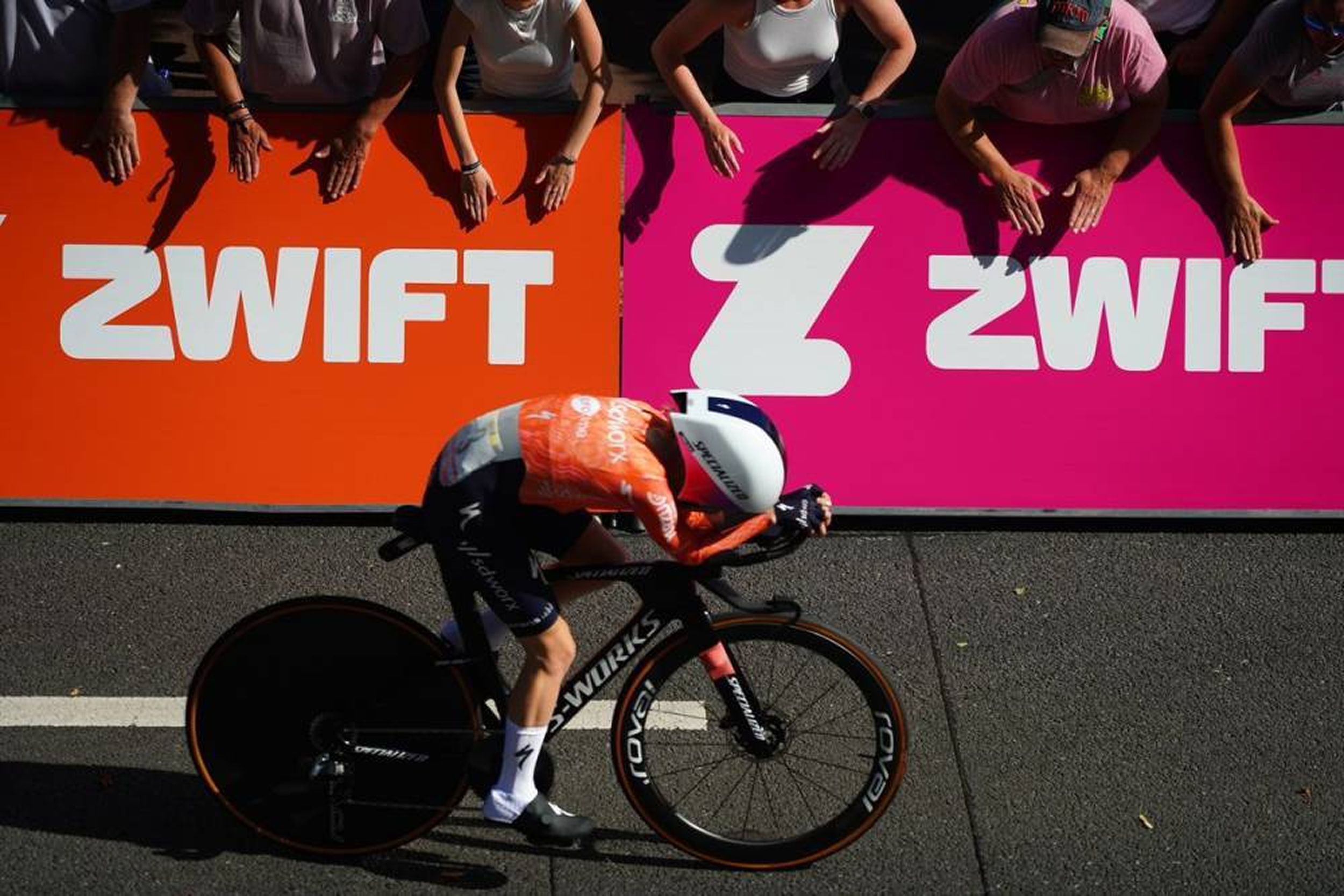 A cyclist in orange and black gear riding a black Specialized bike with deep-dish wheels, in front of a vibrant pink and orange ZWIFT promotional banners, with spectators' hands reaching toward the banners.