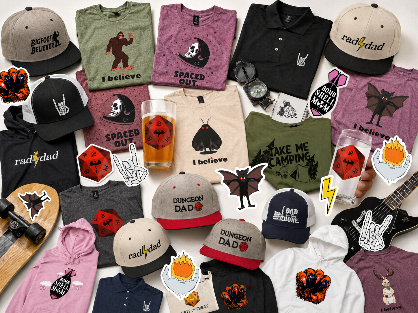 A collection of clothing, hats, stickers, and drink glasses arranged on a table, themed around camping, horror, and gaming. Items include T-shirts with slogans like "I believe" and "Take me camping", hats with sayings like "Bigfoot Believer" and "Dungeon Dad", stickers of bats and flames, red dice, and items like a skateboard, watches, and a guitar.