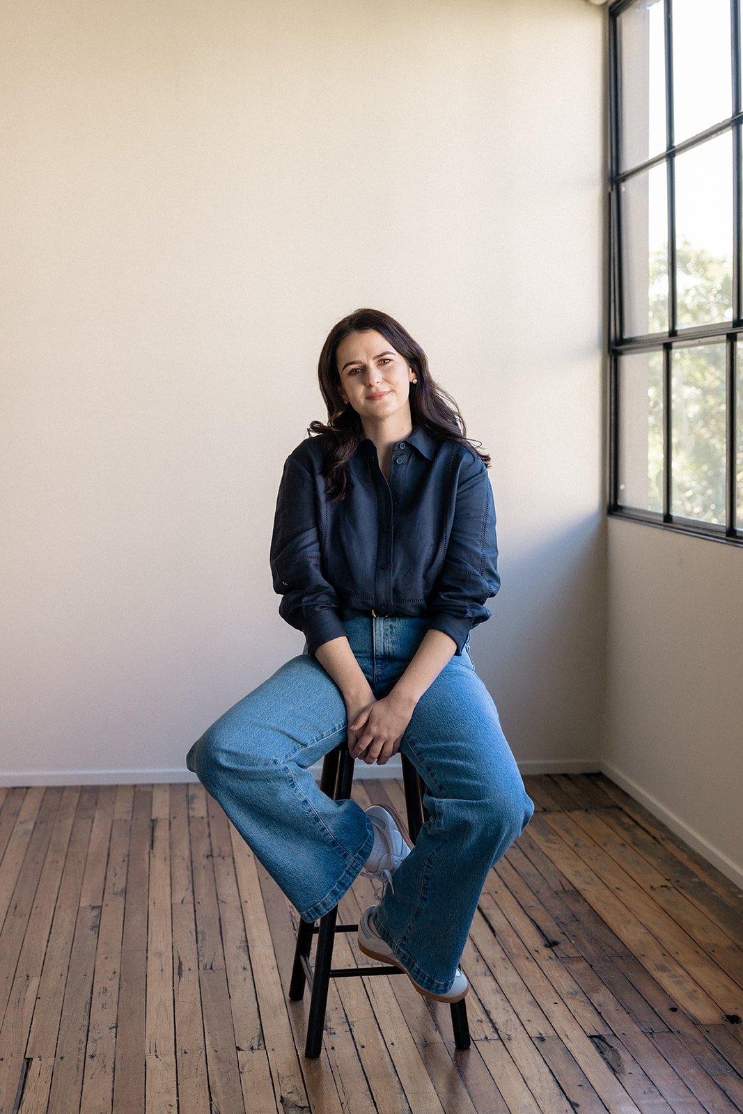 A woman with dark hair in a loose wave, wearing a navy blue button-up shirt, light blue jeans, and white shoes, sitting on a black stool in a room with wooden floors and large window, looking at the camera.