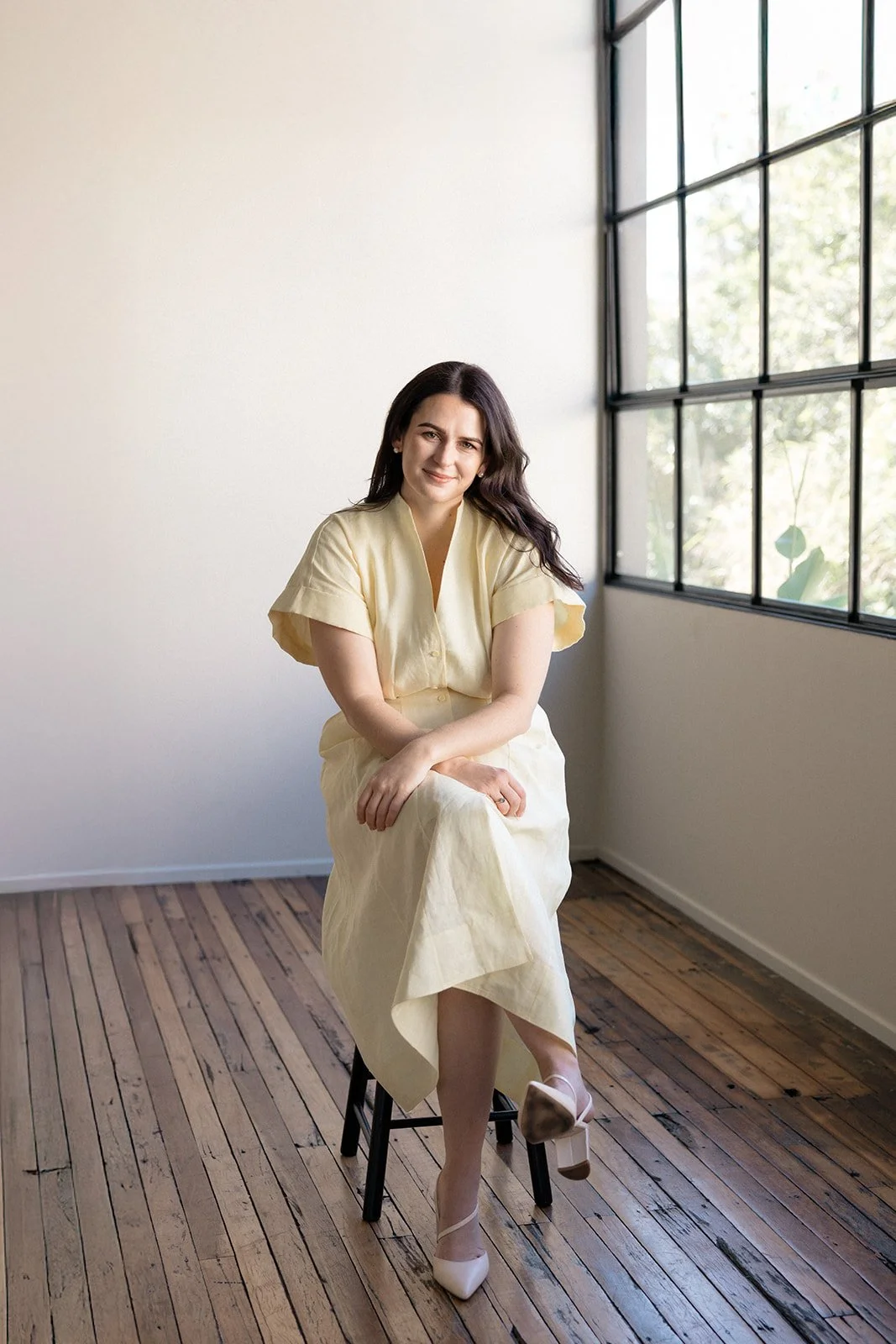A woman with dark hair seated on a black stool near a large window with black frames in a minimally decorated room, wearing a light yellow dress and beige heels.