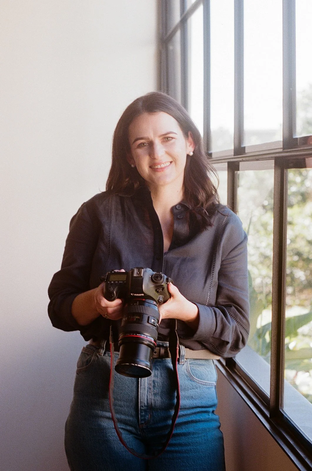 A woman with dark hair holding a Canon camera, standing by large window, smiling at the camera.