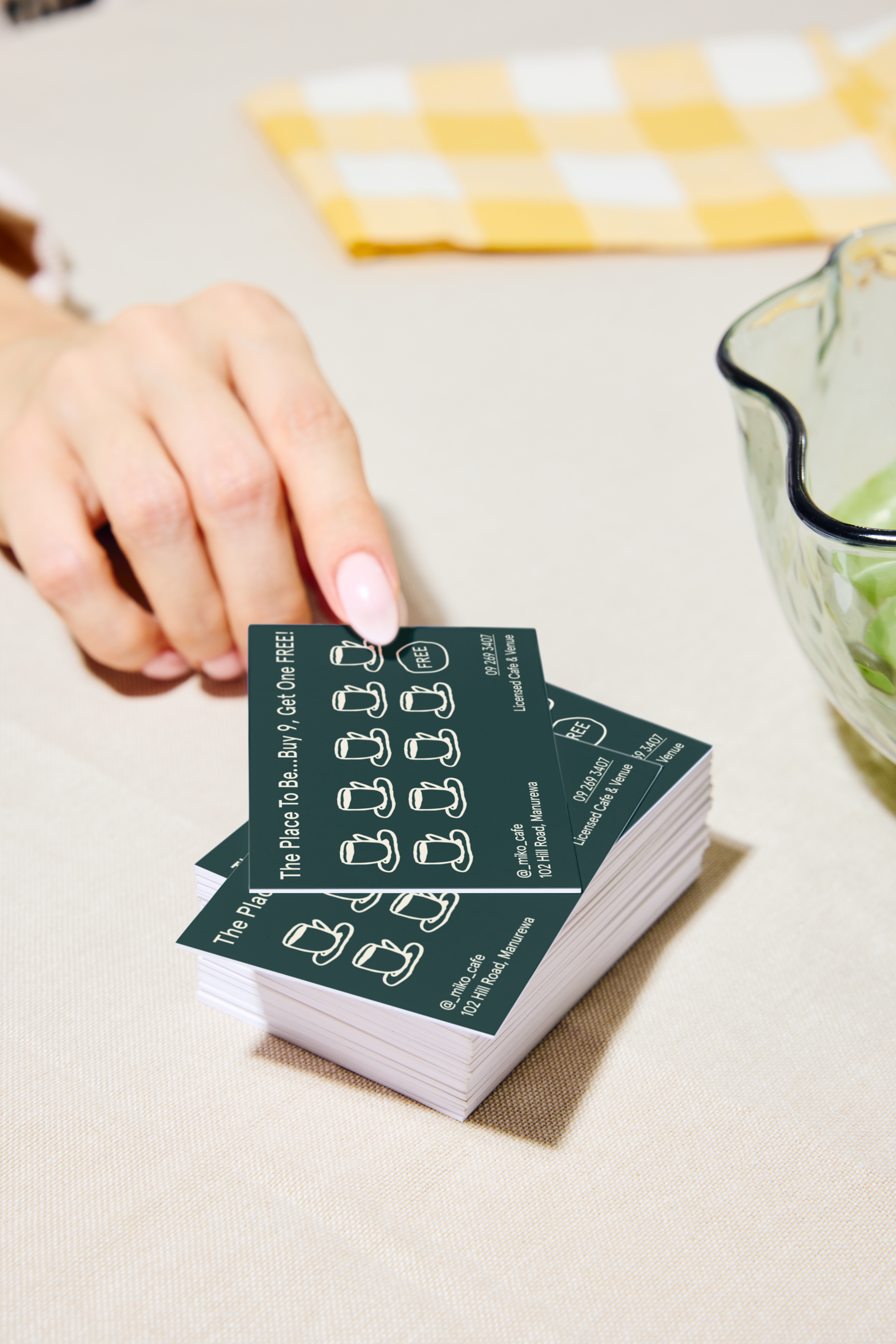 A person holding a stack of dark-colored loyalty or gift cards with white text and icons, placed on a light-colored table, with a yellow-and-white checkered napkin and a partial view of a glass bowl with green food or garnish.