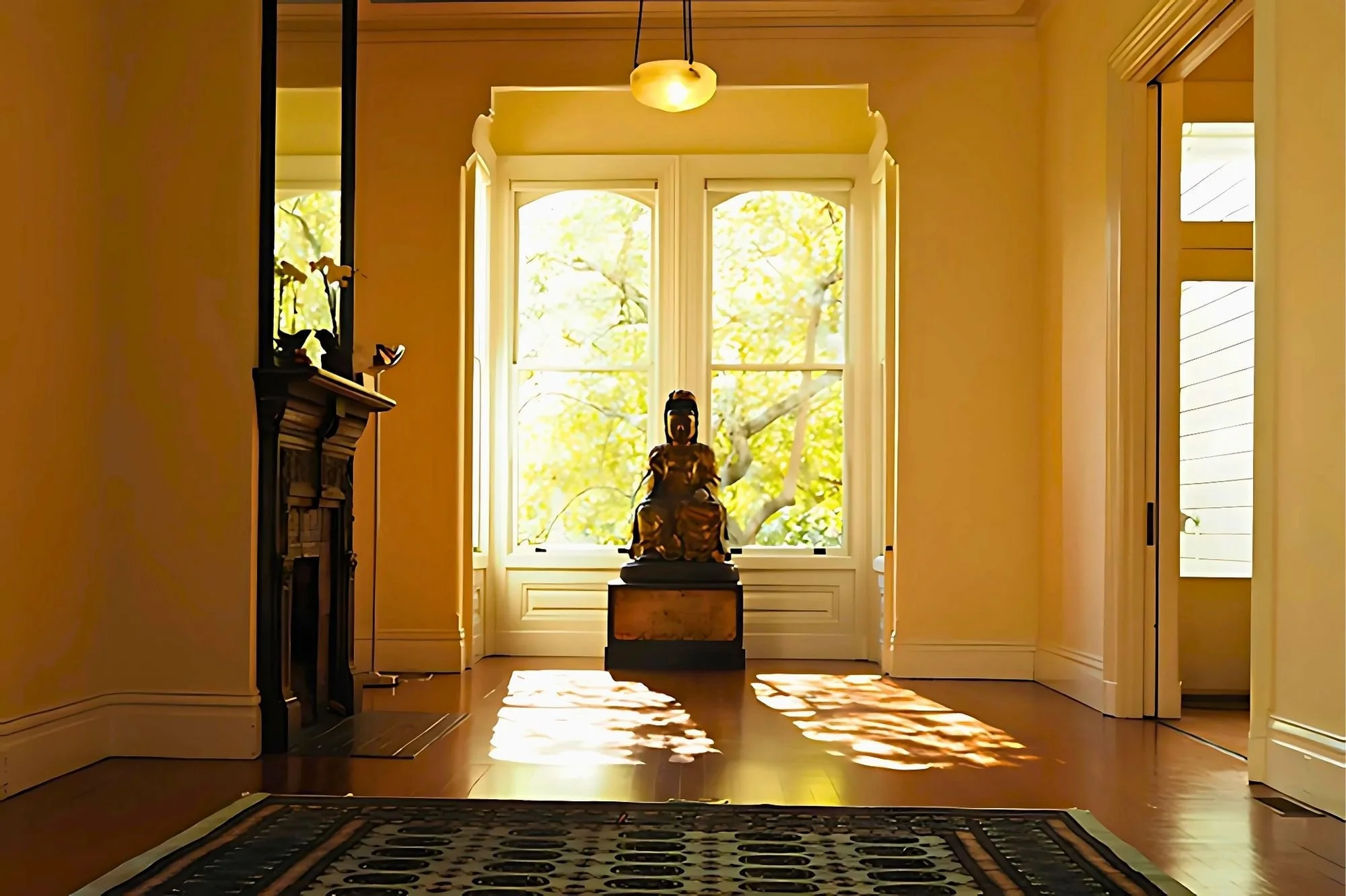 Sunlight streaming through large double-hung windows illuminating a zen sculpture on a pedestal inside a room with wooden floor and patterned rug.