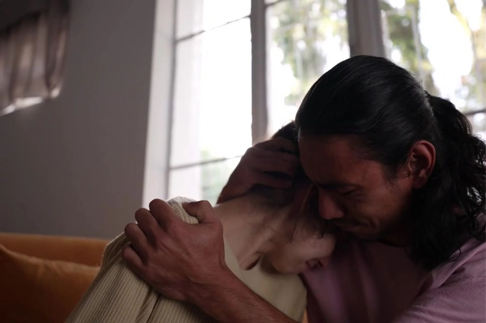A man holding and emotionally comforting his partner in a warm embrace inside a room with natural light coming through the windows.