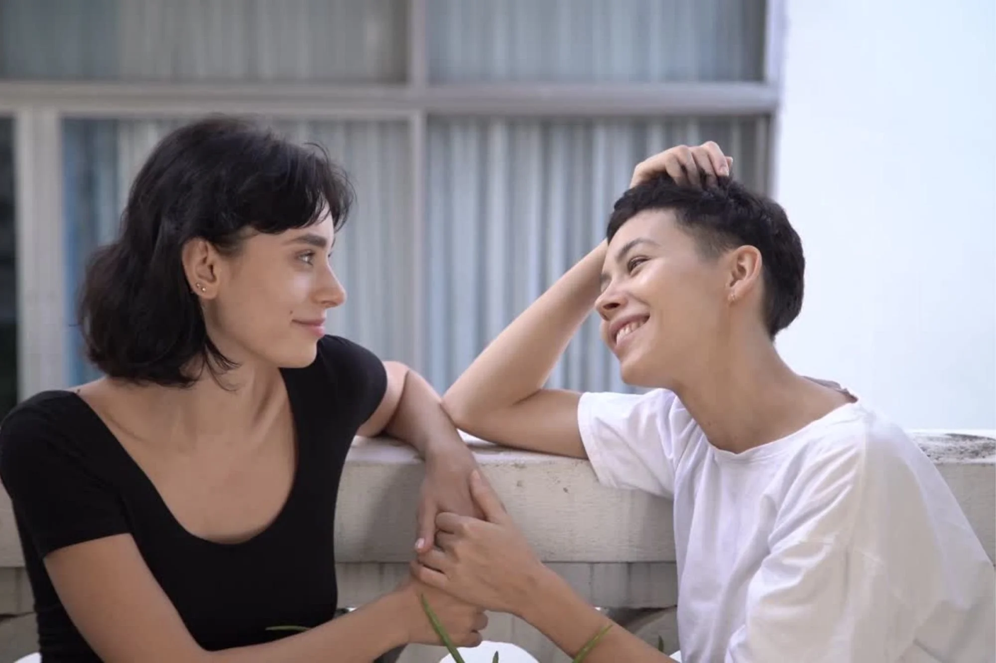 Two women smiling at each other in an indoor setting, with one woman touching the other's head and holding her hand, expressing a moment of connection and affection.