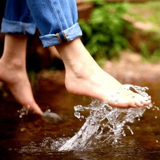 Person with rolled-up jeans splashing in a shallow stream with greenery in the background.
