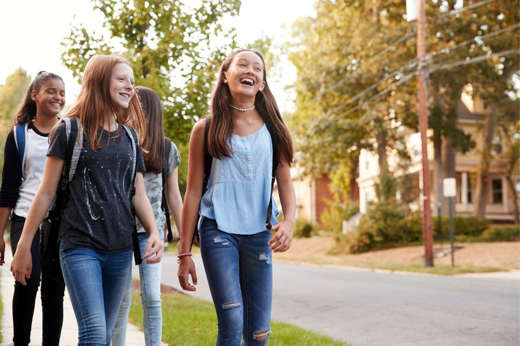 Group of five middle-school girls walking outdoors on a sunny day, smiling and talking, with trees and residential houses in the background.