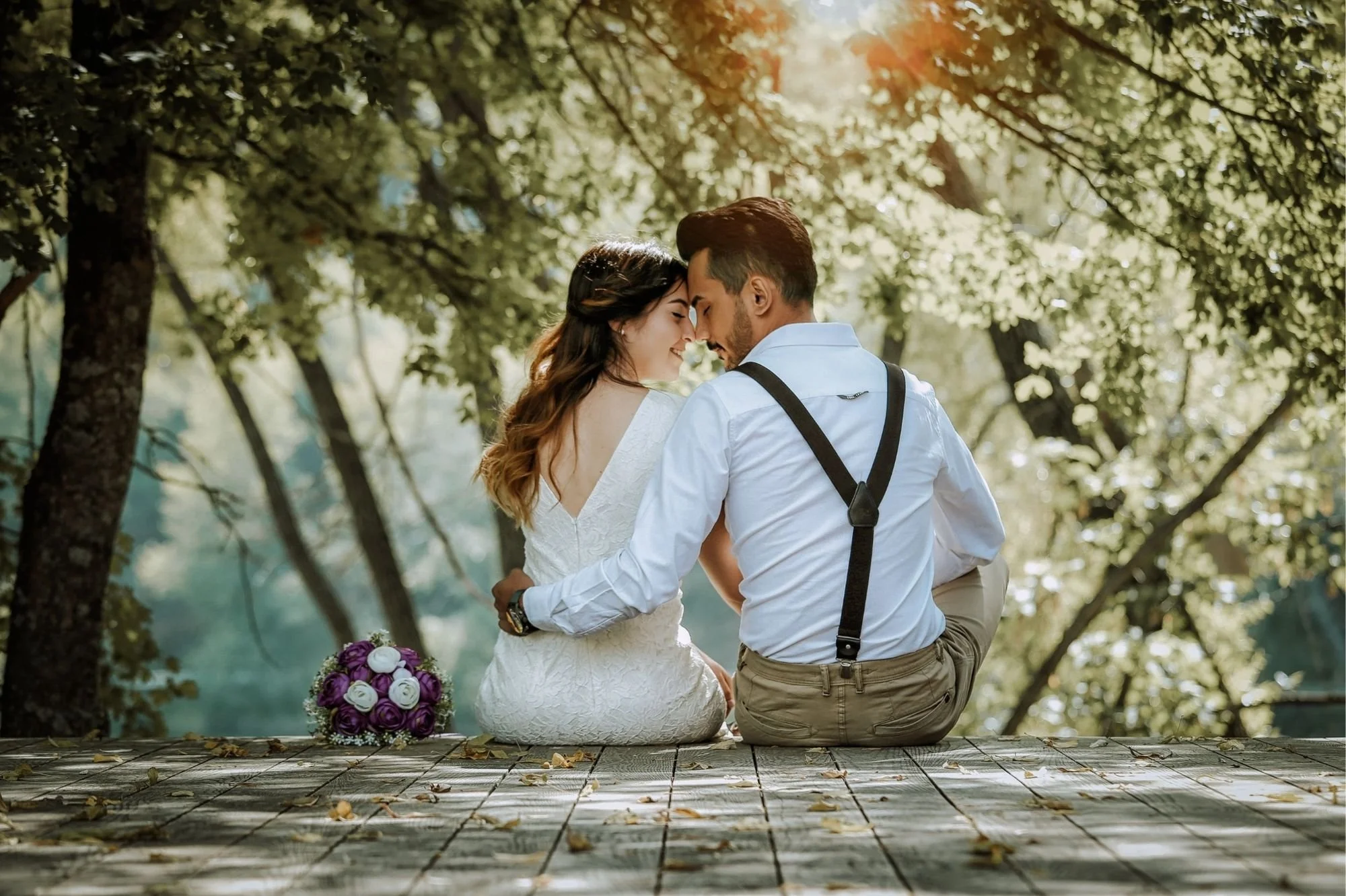 A couple sitting closely together outdoors on a wooden platform, with the woman in a white dress and the man in suspenders and light shirt, surrounded by trees and sunlight, with a bouquet of purple and white roses on the platform.