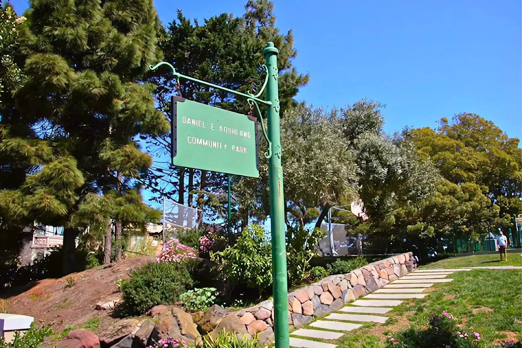 Sign in a park with trees and flowers, reading 'Community Park', with a pathway and a person in the background.
