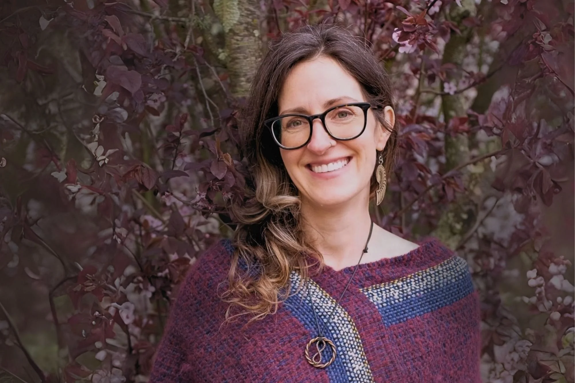 A woman with long, wavy brown hair, glasses, and earrings, smiling in front of a bush with dark purple leaves. She is wearing a multicolored, textured sweater and a necklace with a circular pendant.