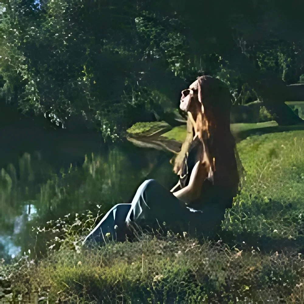 A middle-school girl takes in a reflective moment under moonlight, sitting by a pond in a grassy area surrounded by trees.