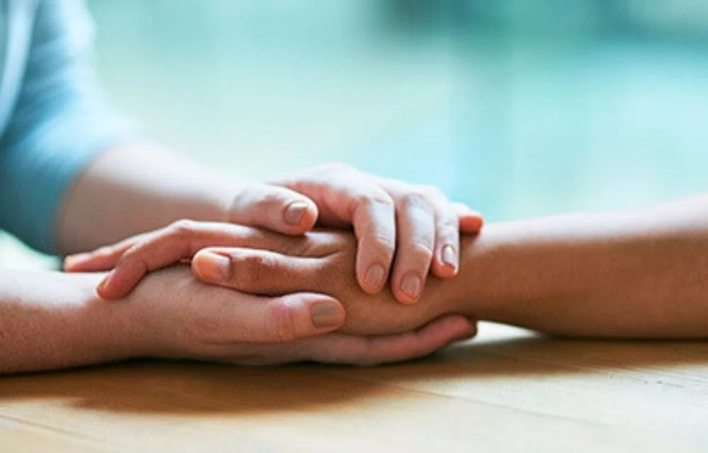 Two pairs of hands, one resting gently on top of the other, on a wooden table.