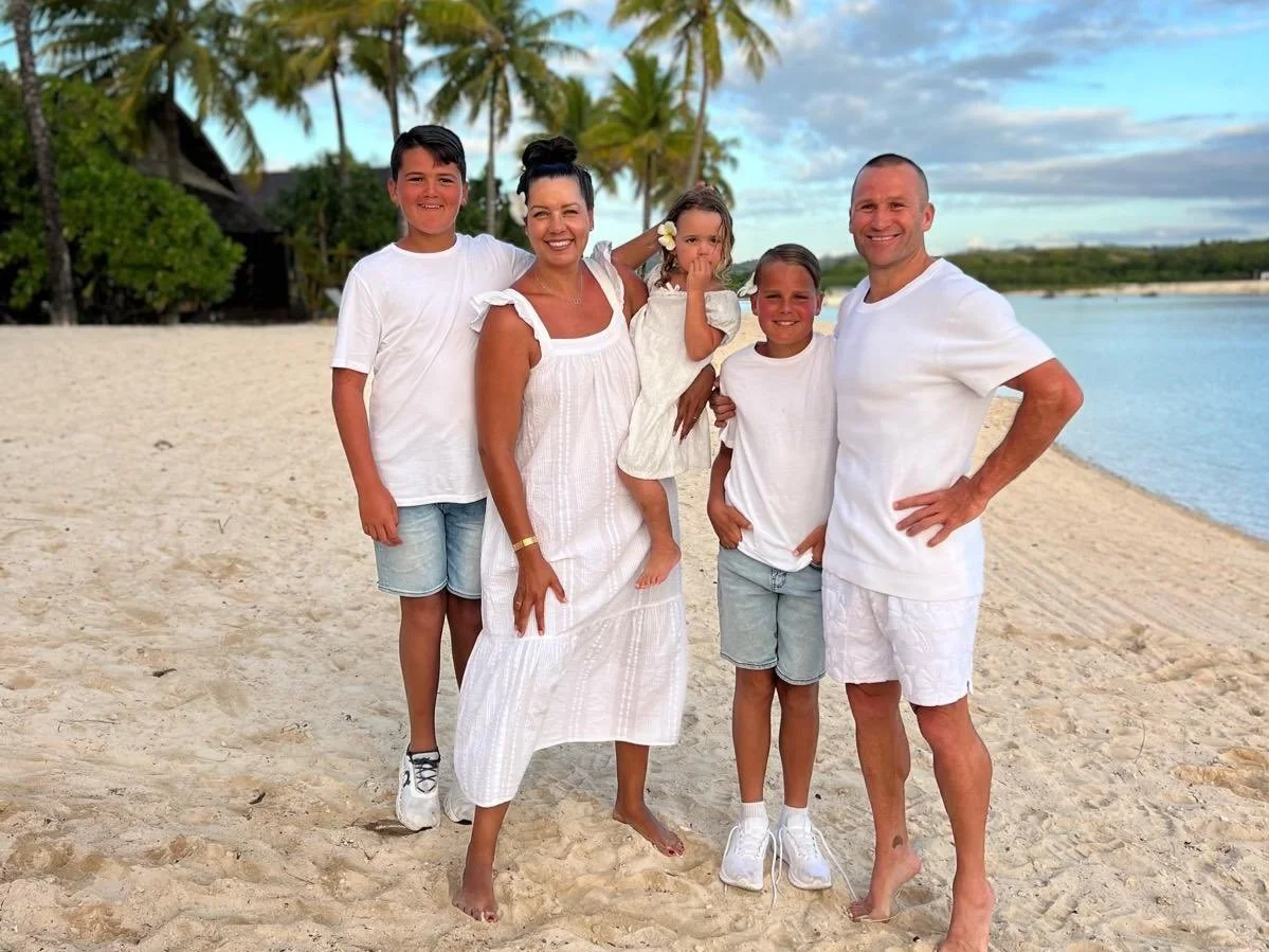 A family of five stands on a sandy beach with palm trees and water in the background, smiling at the camera.