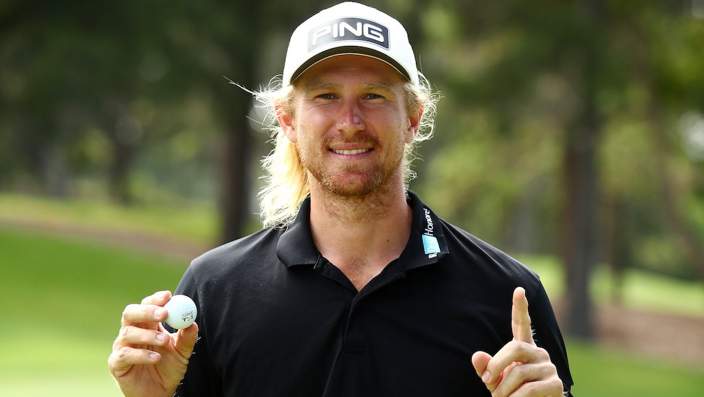 A man with long blonde hair, a beard, wearing a white PING golf cap and black polo shirt, holding a golf ball and making a gesture with his index finger, standing on a golf course.