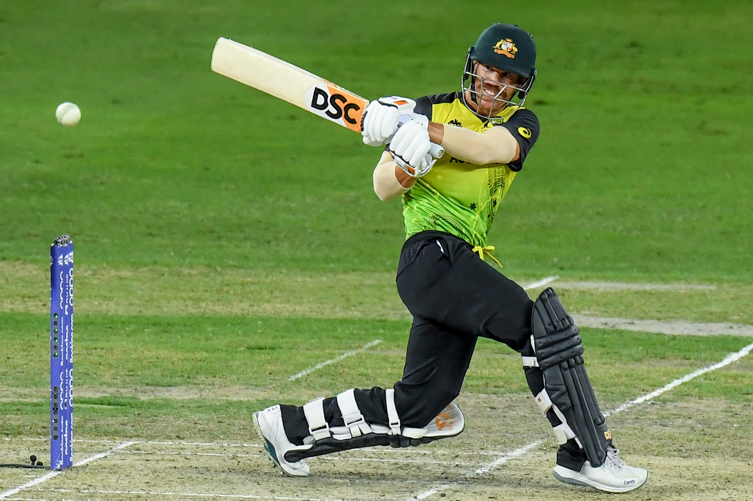 Cricket player in yellow and black uniform swinging a bat at a ball during a match.