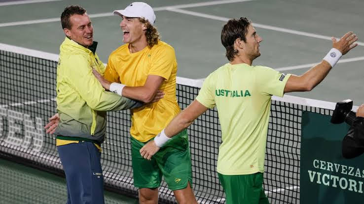 Two male tennis players in yellow and green uniforms celebrating on a tennis court, with one player patting the other on the back and another player walking away.