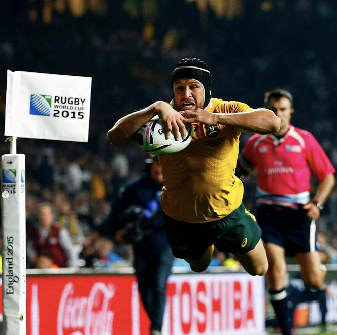 A rugby player in a yellow jersey and black headgear is diving forward and holding a rugby ball during a game at the 2015 Rugby World Cup, with a referee in a pink uniform and spectators in the background.