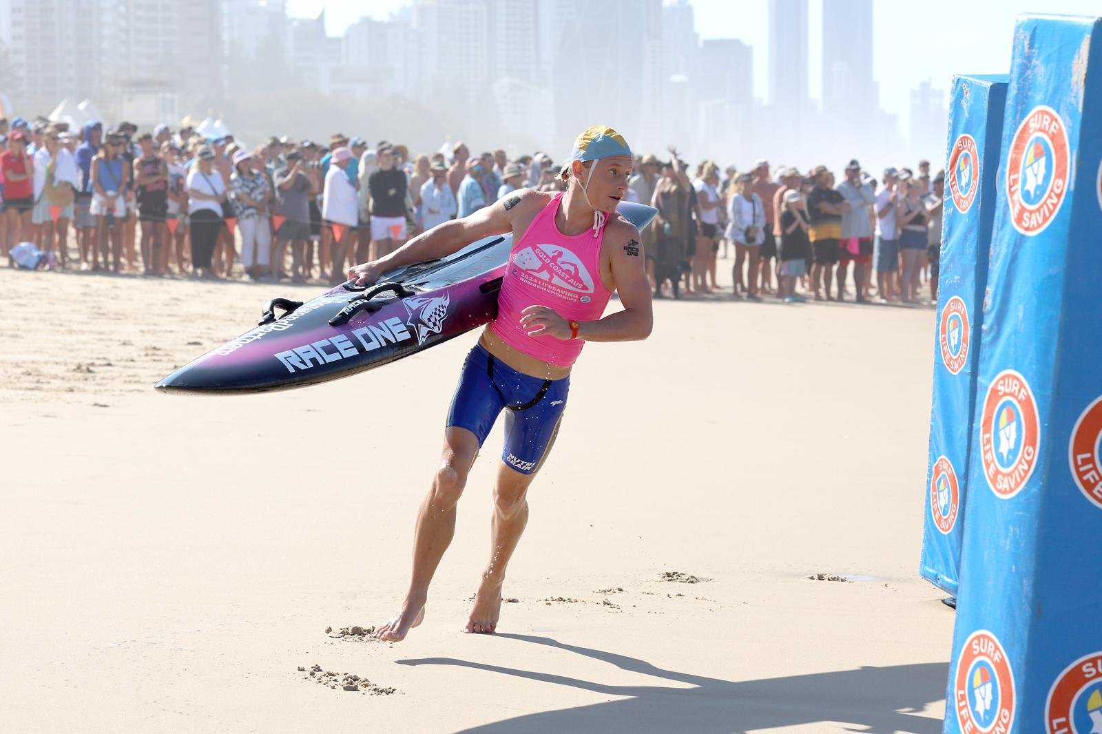 A female surfer carrying a surfboard on a sandy beach during a competition, with spectators in the background.