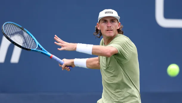 Male tennis player in a green shirt and white cap preparing to hit a tennis ball with a blue racket during a match.