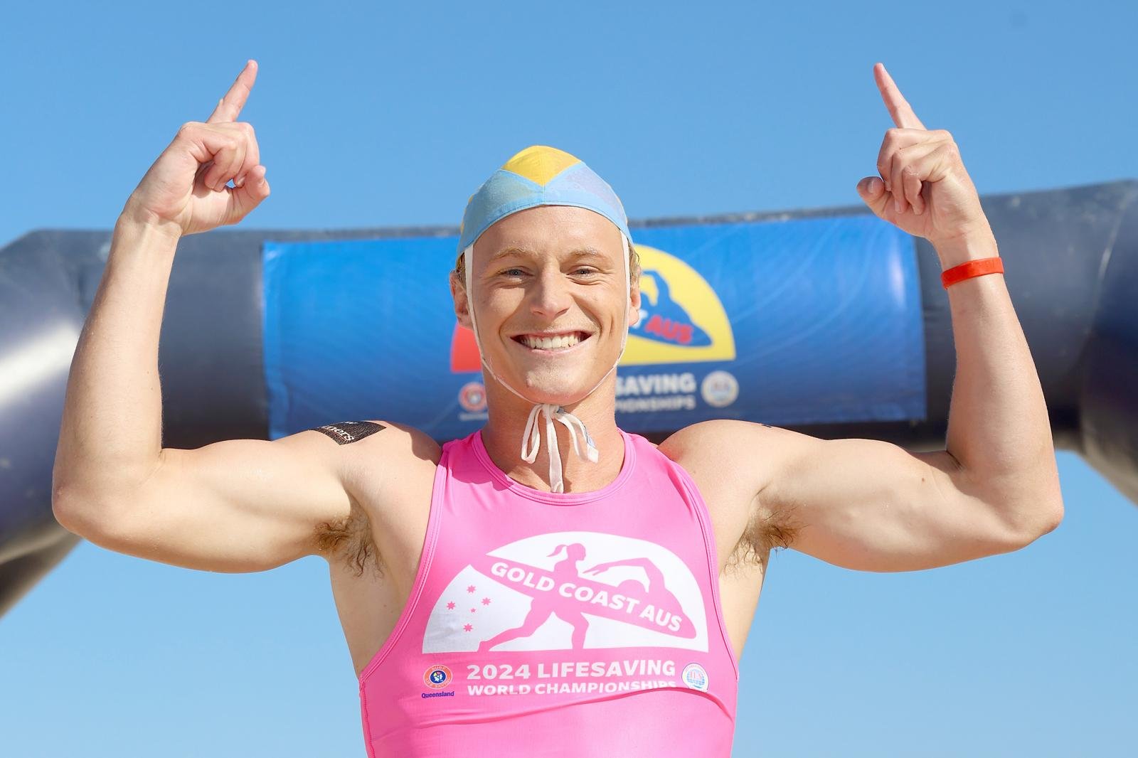 A smiling man in a pink 2024 Lifesaving World Championships shirt, flexing muscles, wearing a blue swim cap, standing in front of a Lifesaving Australia banner on a sunny day.