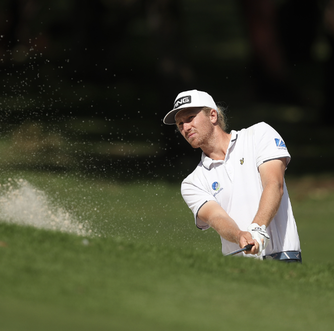 A male golfer in a white shirt and cap swings a golf club out of a bunker, with sand flying in the air.
