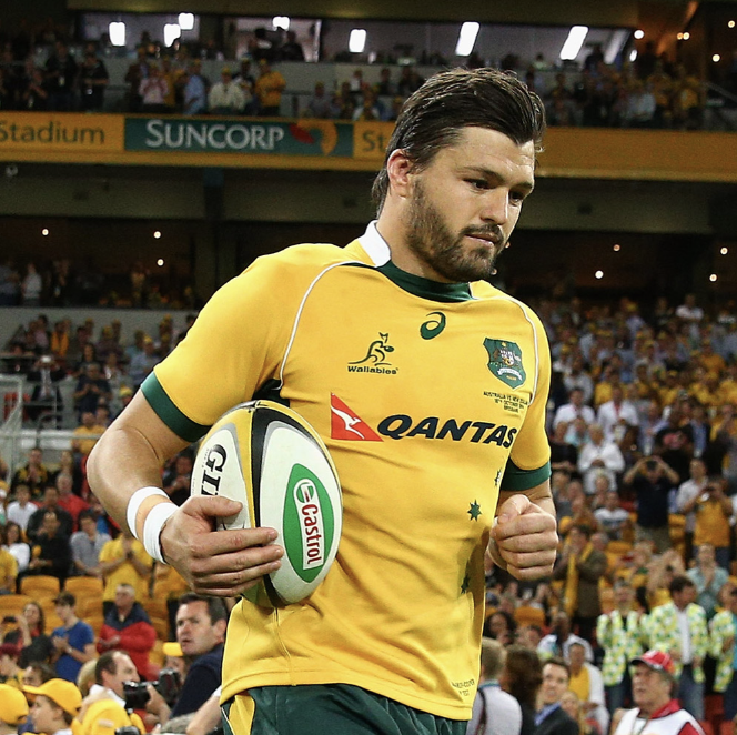 A rugby player wearing a yellow and green Australia team jersey holding a rugby ball on a field with spectators in the background.