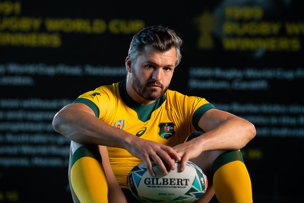 A male rugby player in a yellow and green uniform sitting and holding a rugby ball in front of a dark background with yellow text.