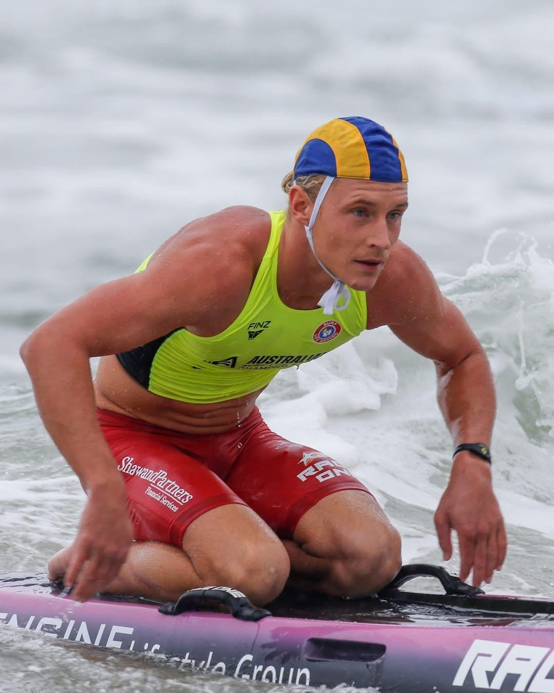 Male athlete in yellow tank top and red shorts, wearing a blue and yellow swim cap, crouched on a paddleboard in the ocean, with water splashing around him.