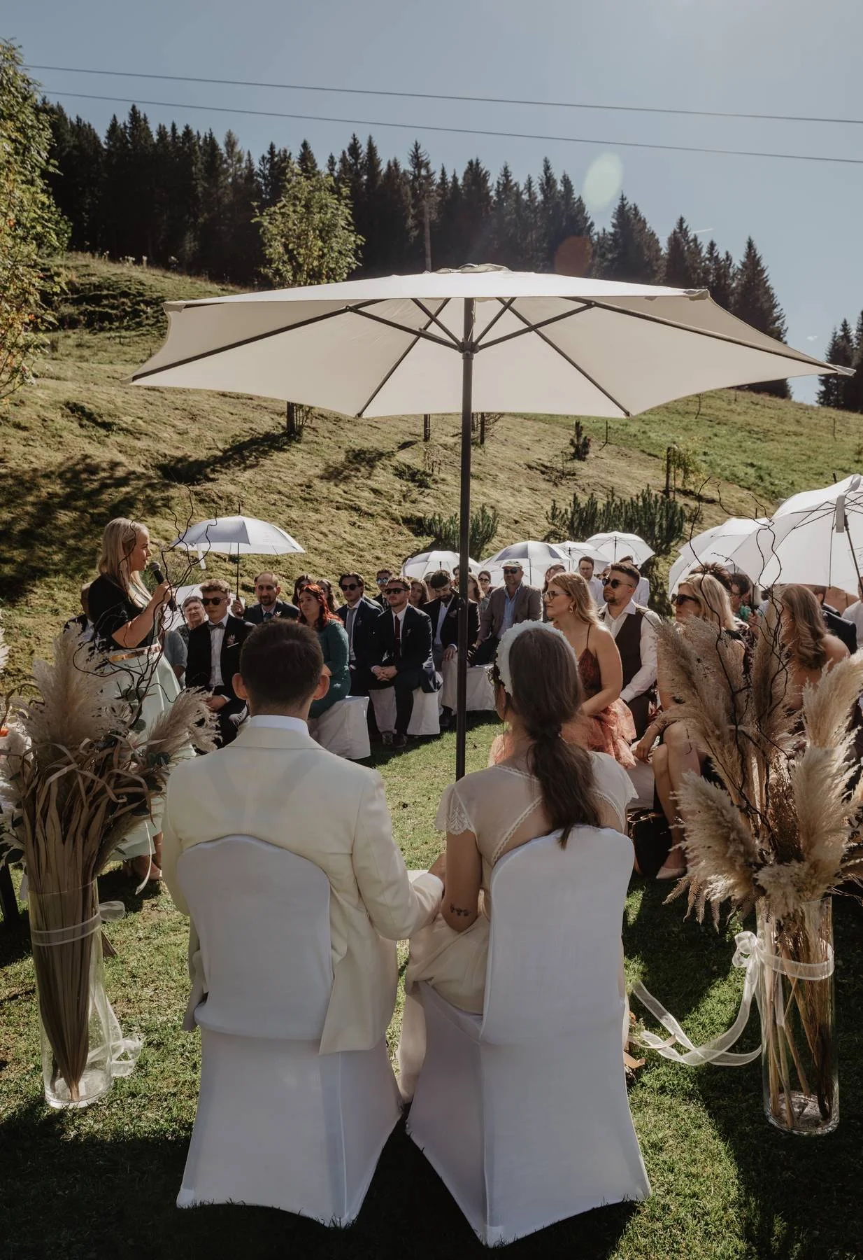 Hochzeit im Freien mit Brautpaar, das sich die Hände hält, während sie vor Gästen sitzen, die unter großen Sonnen-Schirmen auf einer grünen Wiese mit Hügeln und Bäumen im Hintergrund feiern.