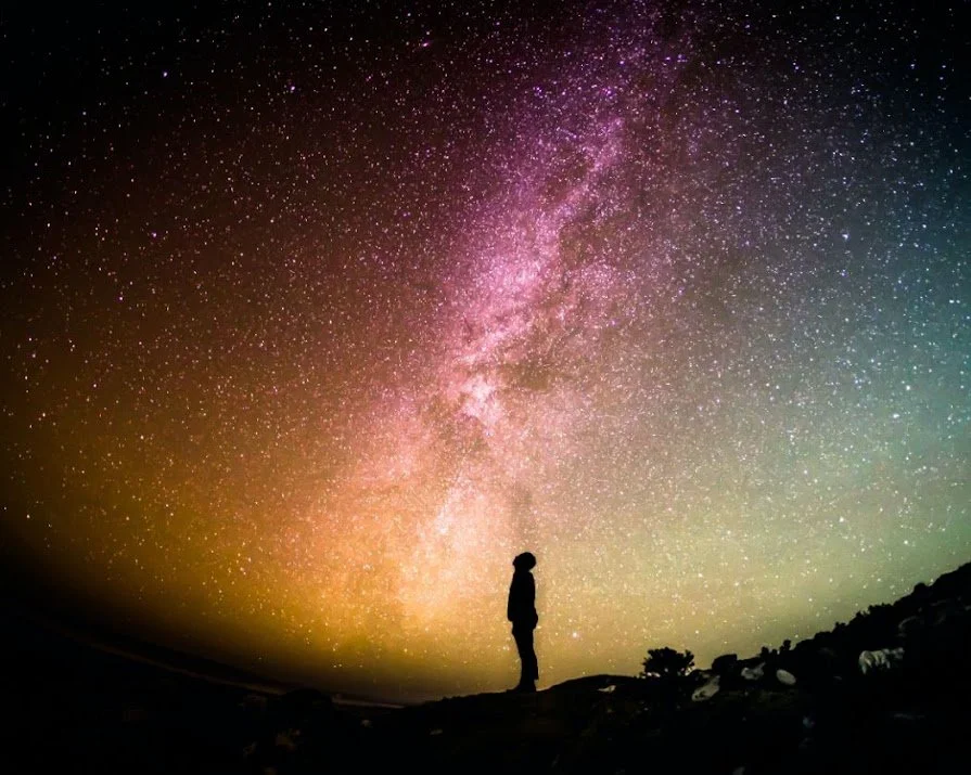 Silhouette of a person standing on a hill under a colorful, star-filled night sky with the Milky Way galaxy prominently displayed.