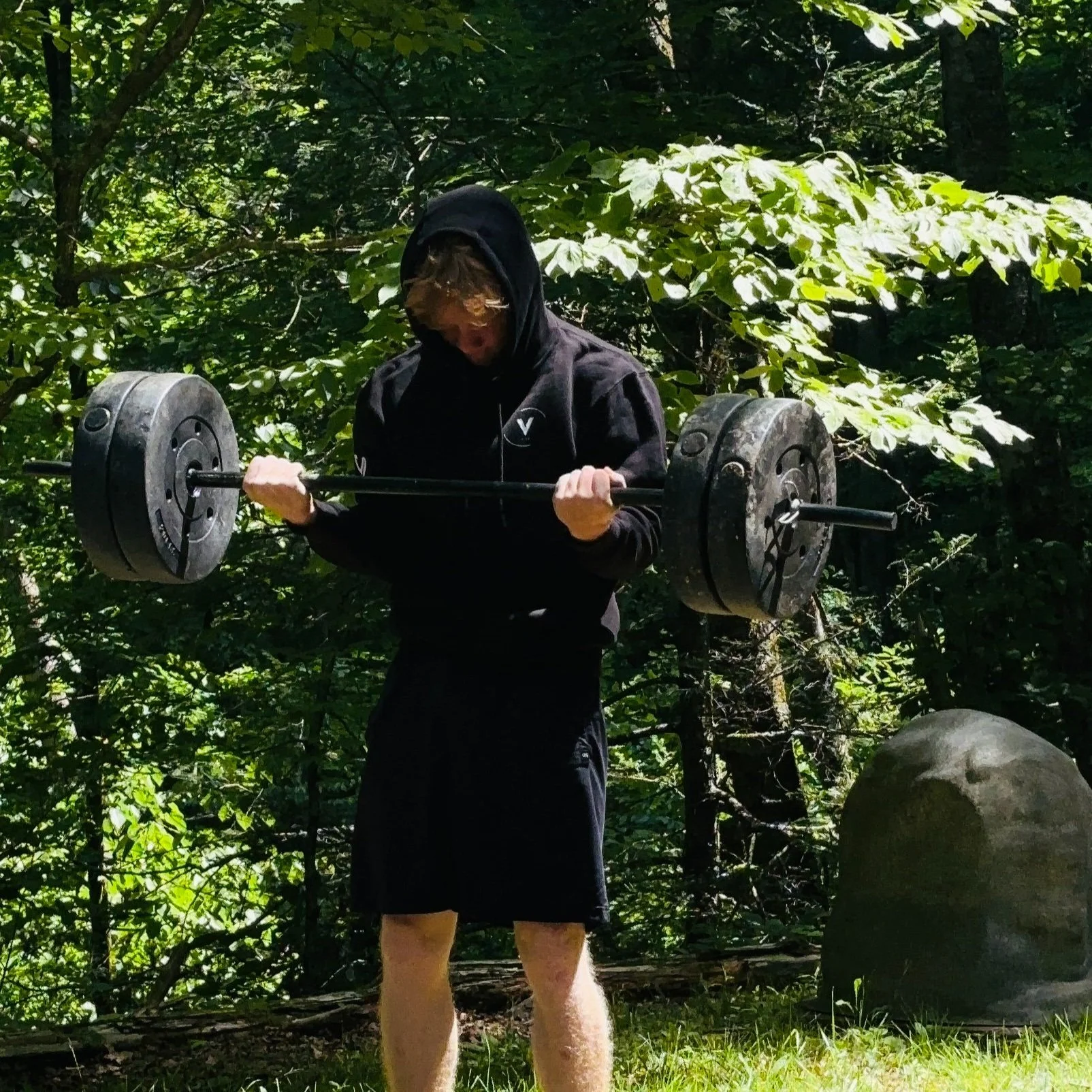 A person in a black hoodie and shorts lifting a barbell with large weights outdoors among green trees and grass.