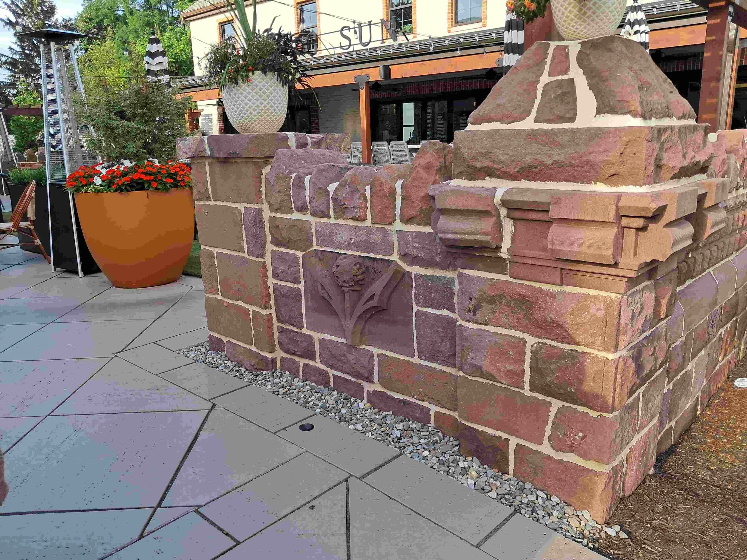 Decorative stone structure with a flower sculpture in the middle, situated on a paved outdoor patio area, adjacent to a large flower pot with red flowers.