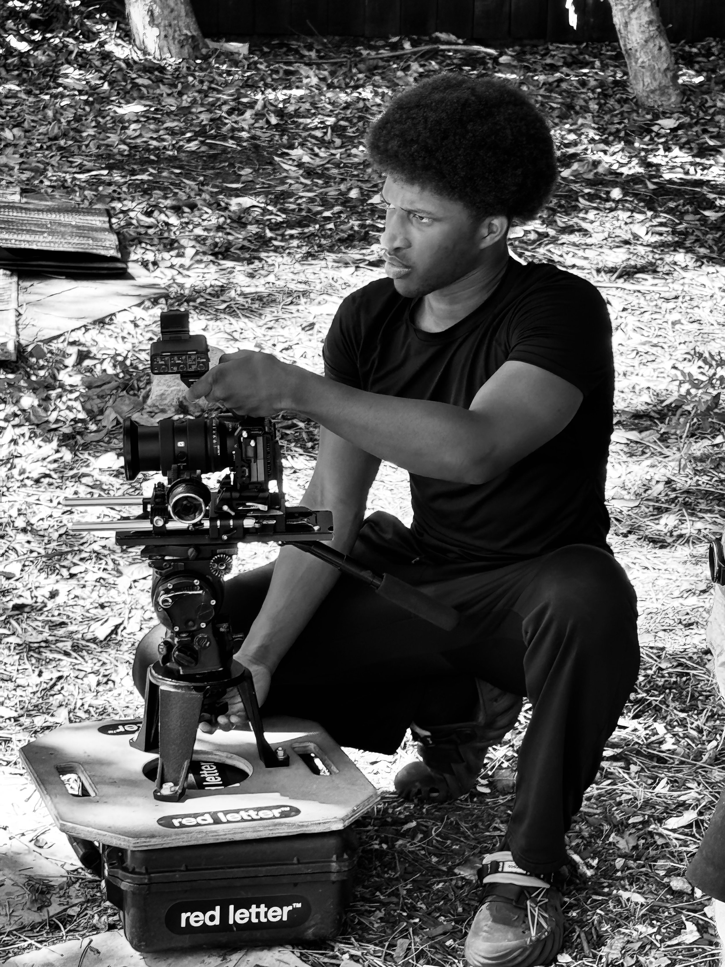 A man with an Afro hairstyle performing camera setup outdoors on leaf-covered ground with trees in background. Lex Lumpkin directing on the set of, "The Death of a Nation"