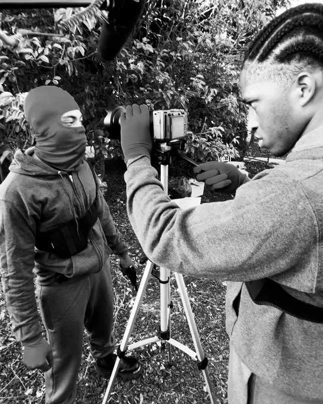 Two young men are filming outdoors, one adjusting a camera on a tripod while the other stands with a covered face, wearing a jacket and gloves, surrounded by trees and foliage. Lex Lumpkin directing Reed Lorenzo Shannon, on set, Dilemma in the Alley.