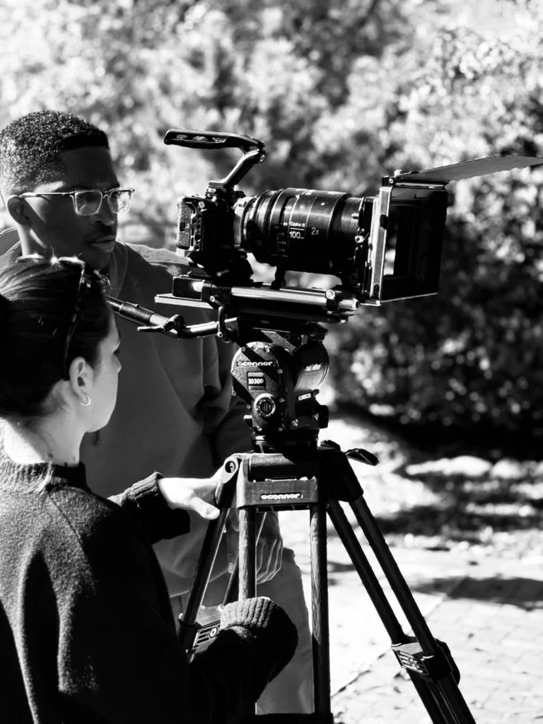 Two people operating a professional video camera outdoors in black and white. Lex Lumpkin and Julia Maixer on the set of The Death of a Nation.