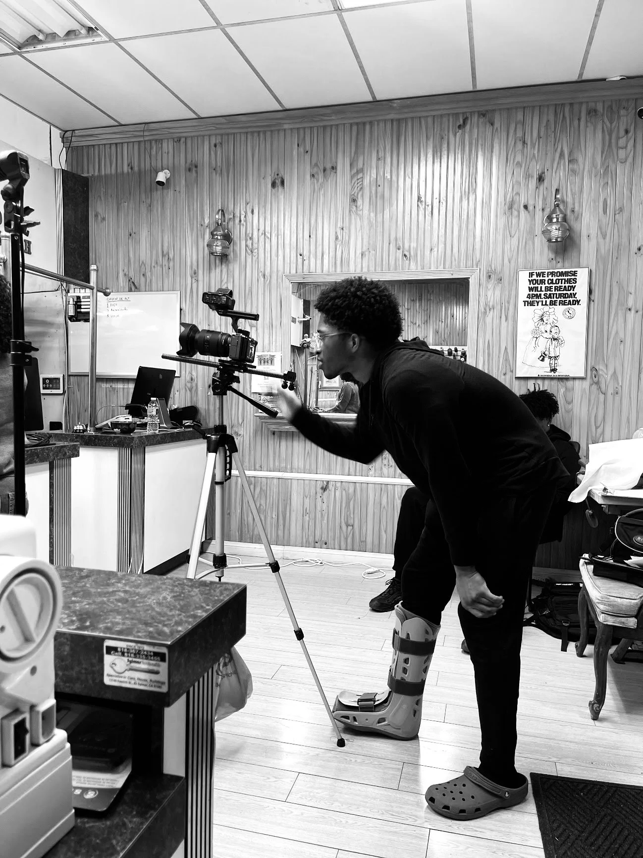 A man with a medical boot on his left foot filming with a camera mounted on a tripod in a room with wooden wall paneling and posters. Lex Lumpkin directing on the set of, "Late Night Pickup"