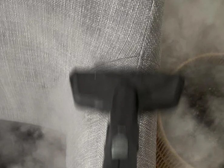 Close-up of a black handheld steam cleaner with steam coming out, being used on a fabric surface.