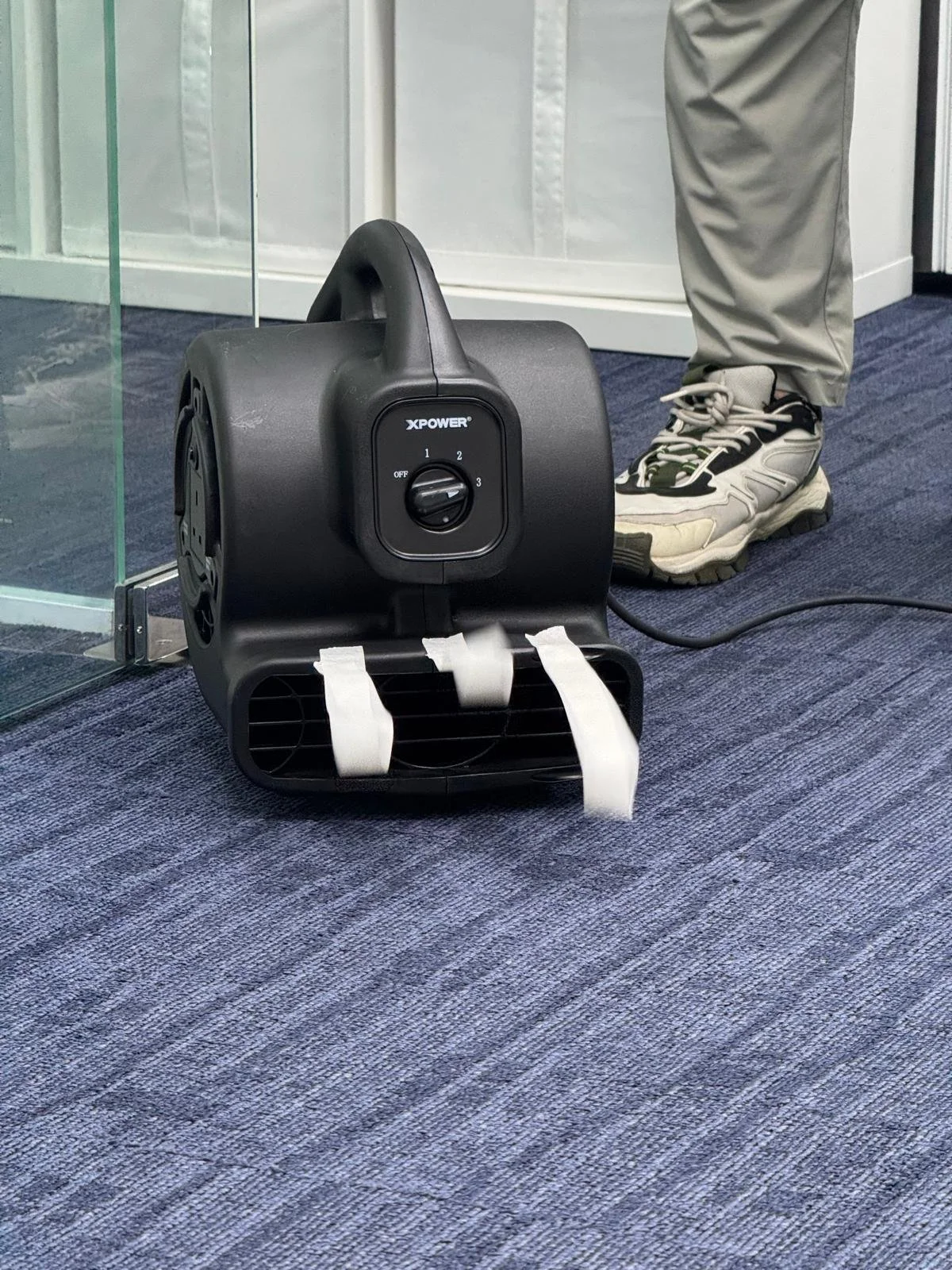 Black portable fan with white tape on its grill, plugged into the nearby power source, with a person's light-colored sneakered foot visible in the background on a blue carpeted floor.