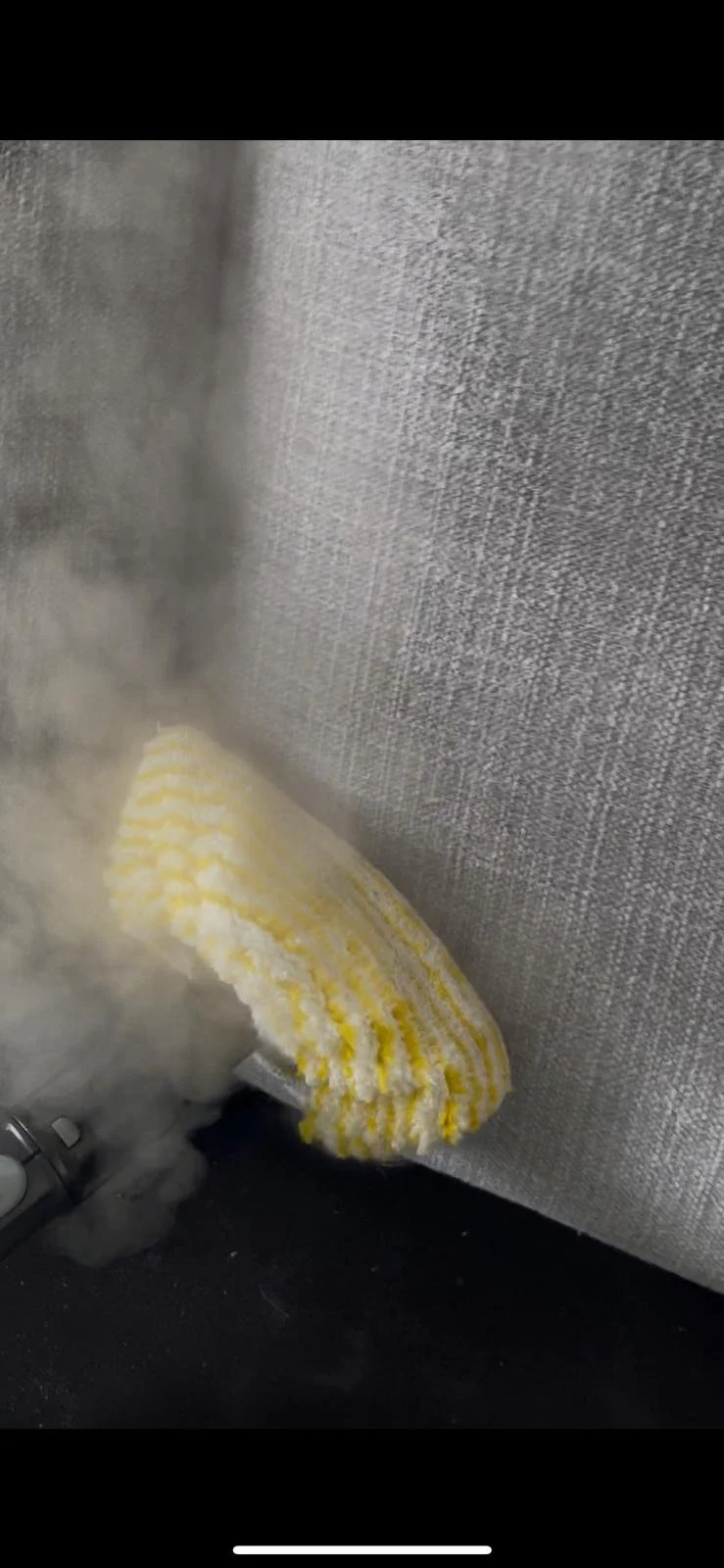 Cleaning a gray upholstered sofa with a yellow and white striped sponge or cloth, with visible steam or cleaning vapor.