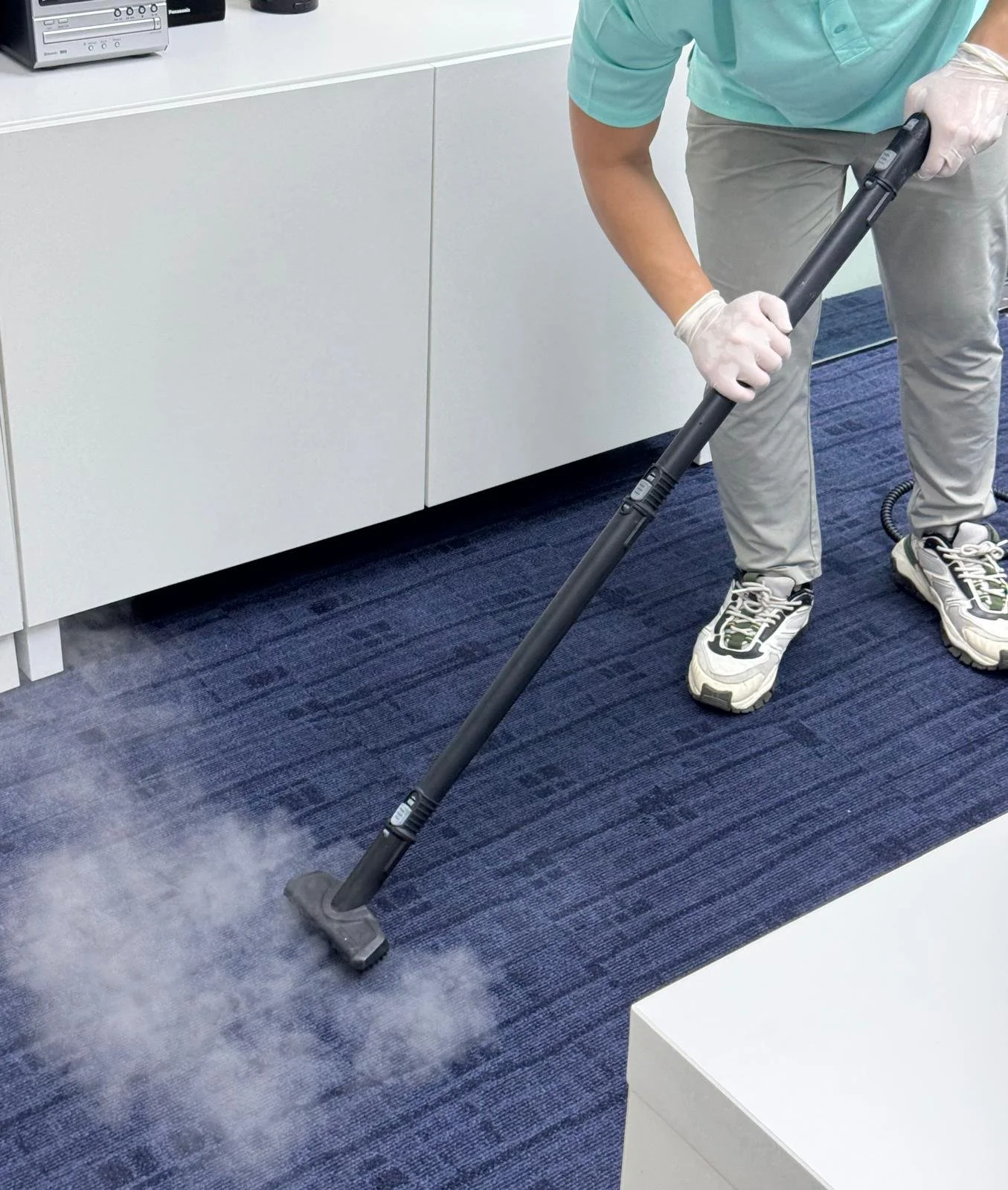 A person wearing gloves and sneakers is using a steam cleaner to disinfect a blue carpet in an office setting.