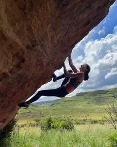 A woman in athletic clothing rock climbing outdoors on a large overhanging cliff with green fields and blue sky in the background.