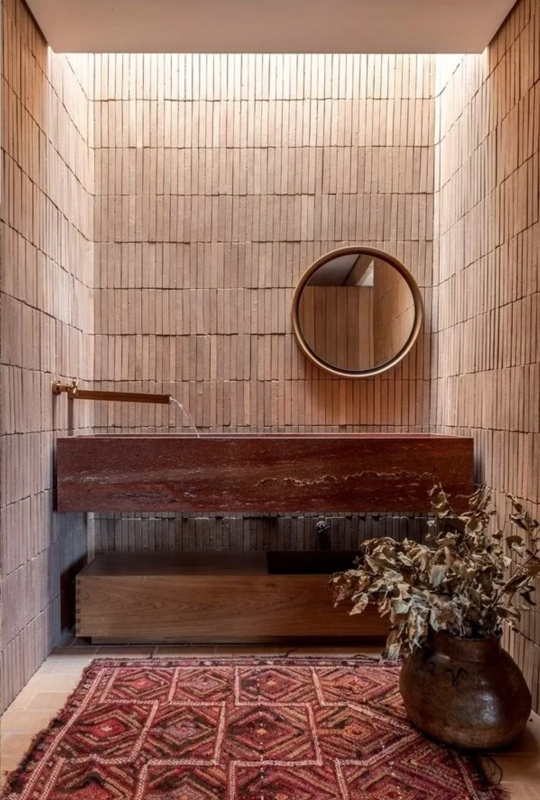 A small bathroom with brick walls, a round mirror, a stone sink, and a vase with dried leaves, a red patterned rug on the floor.