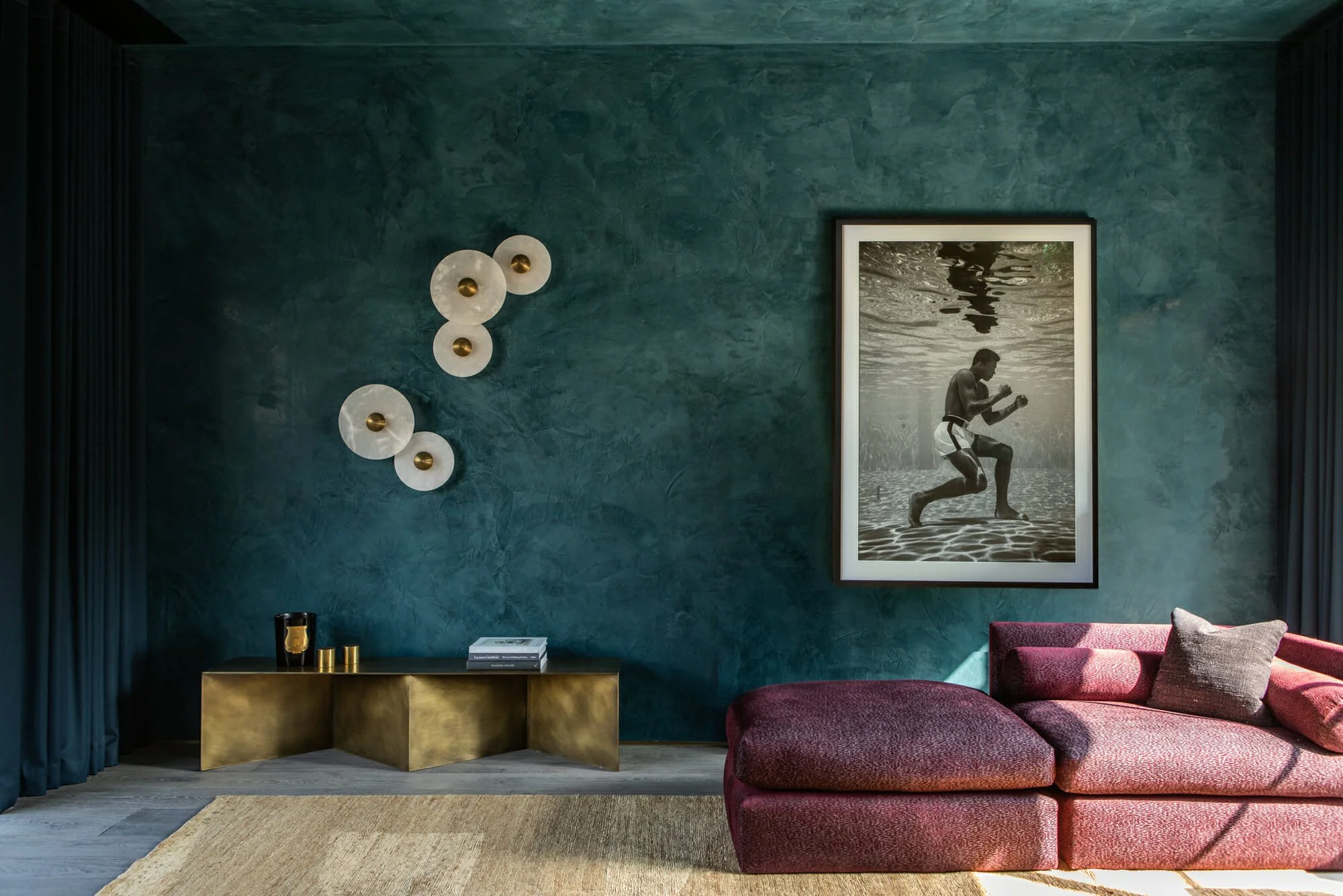 Living room with a textured teal wall, a large black and white framed photograph of a scuba diver, a magenta sofa with cushions, a contemporary gold coffee table, and wall decor with white circular elements with gold centers.