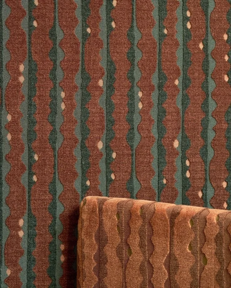 Close-up of a patterned fabric with vertical wavy stripes in brown, green, and beige, with some circular embellishments, and a section of upholstered furniture matching the fabric.