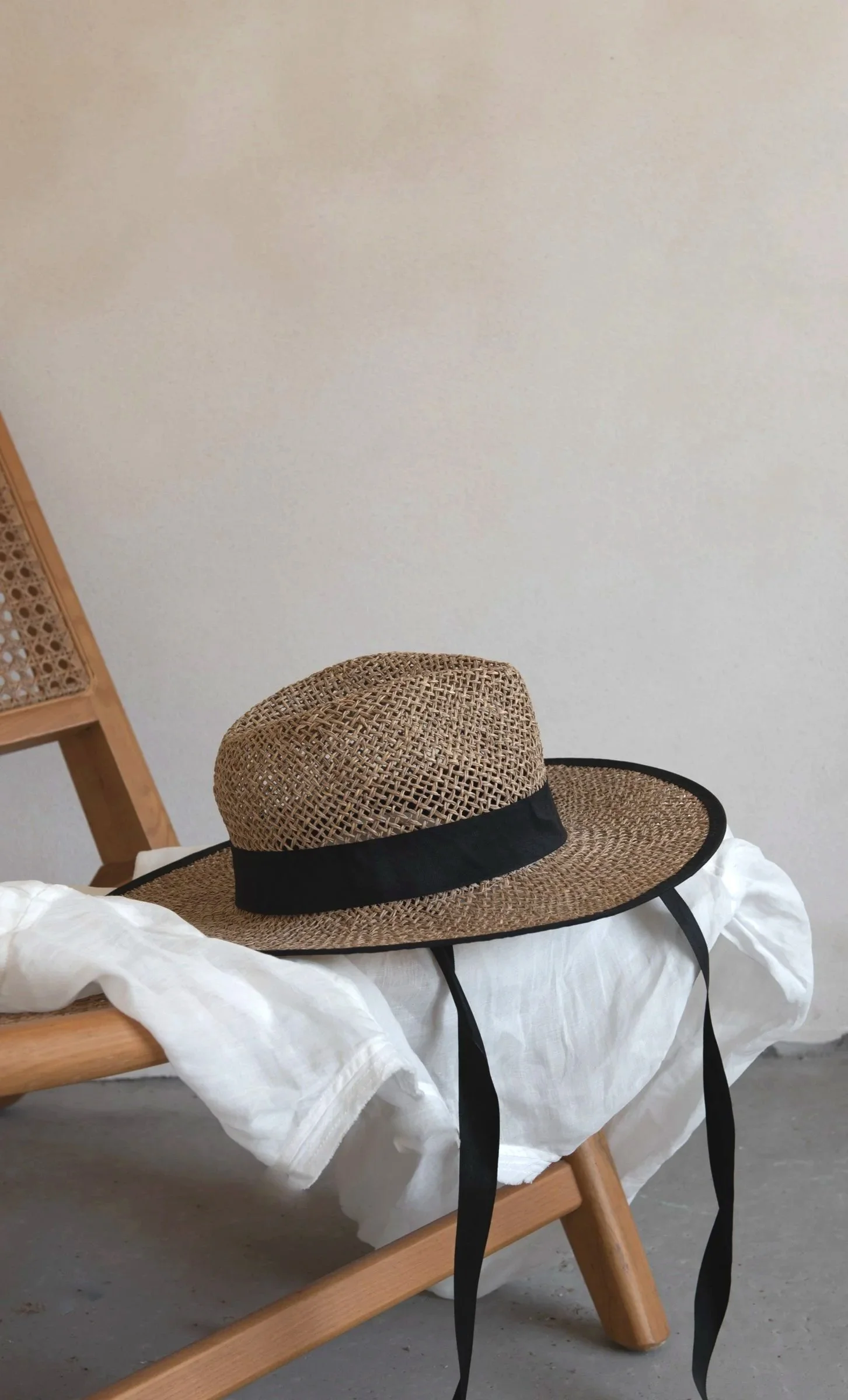 A woven straw hat with a black band and black strings resting on a white cloth-draped wooden chair.
